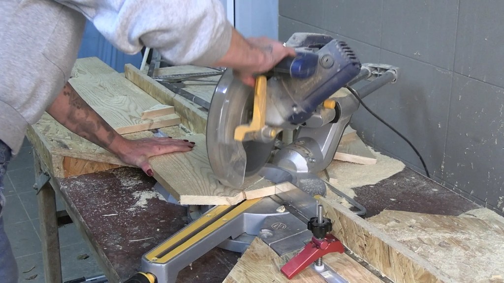 Person sägt ein Holzbrett mit einer Kapp- und Gehrungssäge zu; Holzspäne liegen auf der Werkbank, im Hintergrund weitere Bretter und Werkzeug.