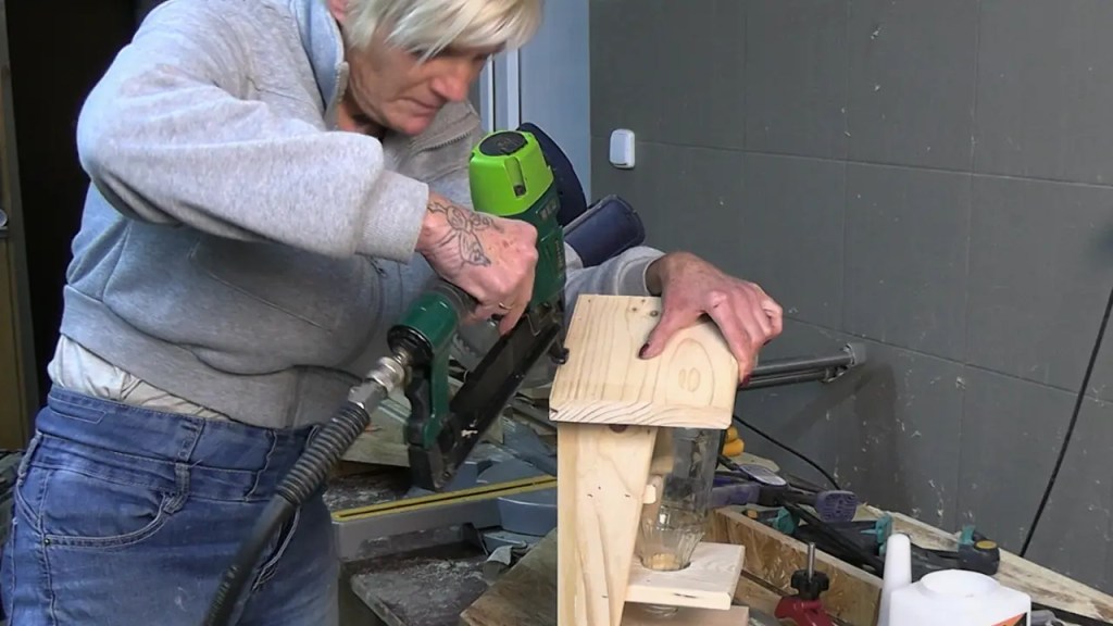 Person befestigt das Dach eines selbstgebauten Vogelfutterhauses mit einem Drucklufttacker; das Holzhaus mit eingesetzter Glasflasche steht auf einer Werkbank in einer kleinen Werkstatt.