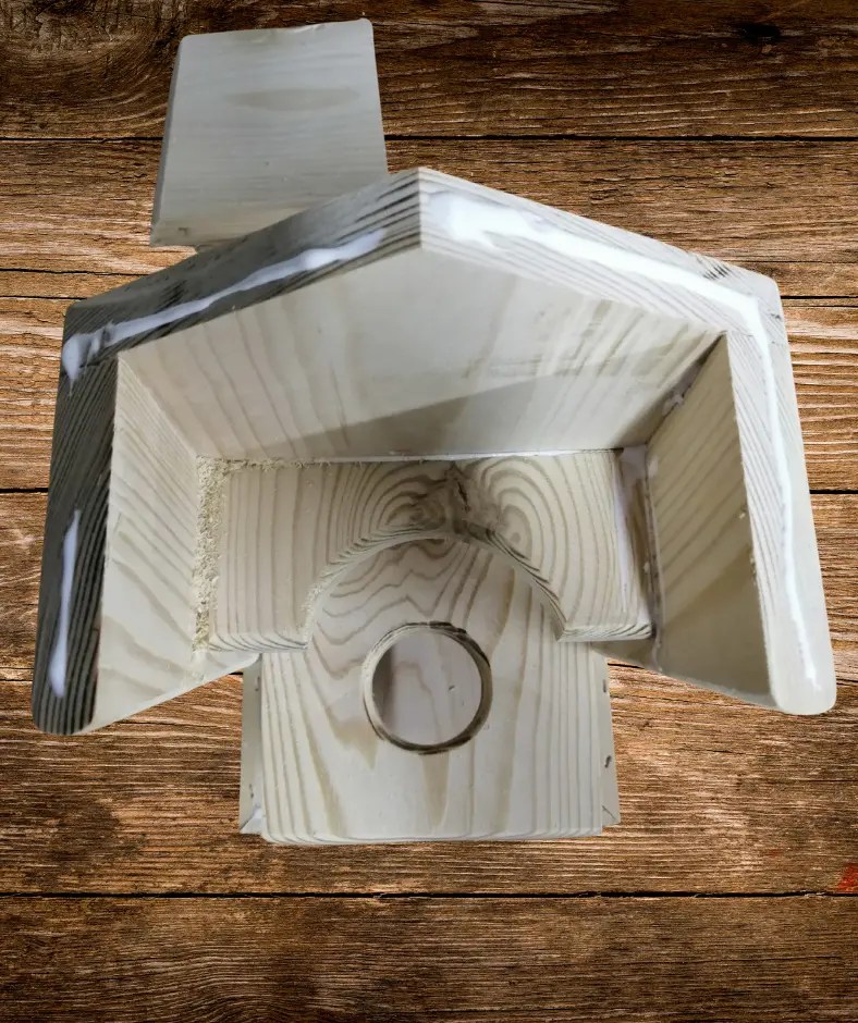 Teilweise zusammengebautes Vogelfutterhaus aus hellem Holz: Rückwand, Seiten und Boden sind montiert, die Leimfugen sind sichtbar; in der Front ist ein rundes Loch für das Futtersilo eingearbeitet.