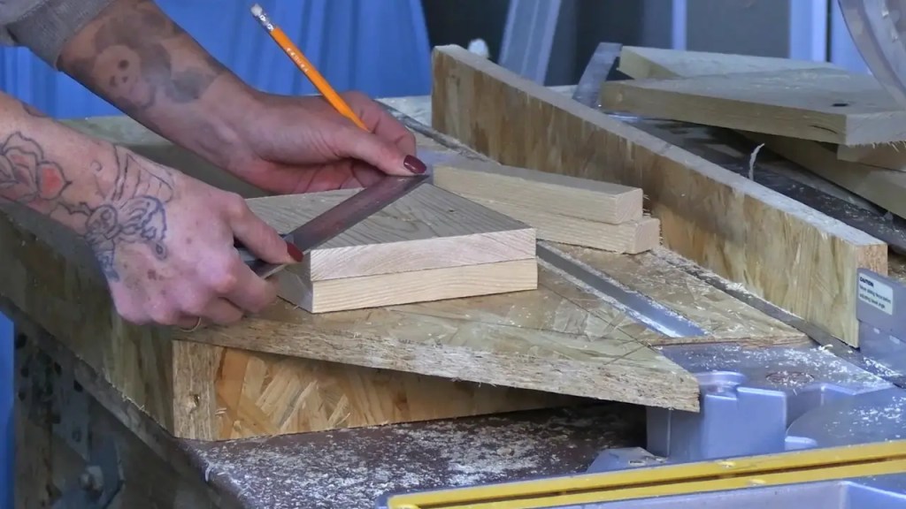 Person markiert eine Schnittlinie auf einem kleinen Holzbrett mit Winkel und Bleistift; weitere zugeschnittene Holzstücke und Sägewerkzeug liegen auf der Werkbank.