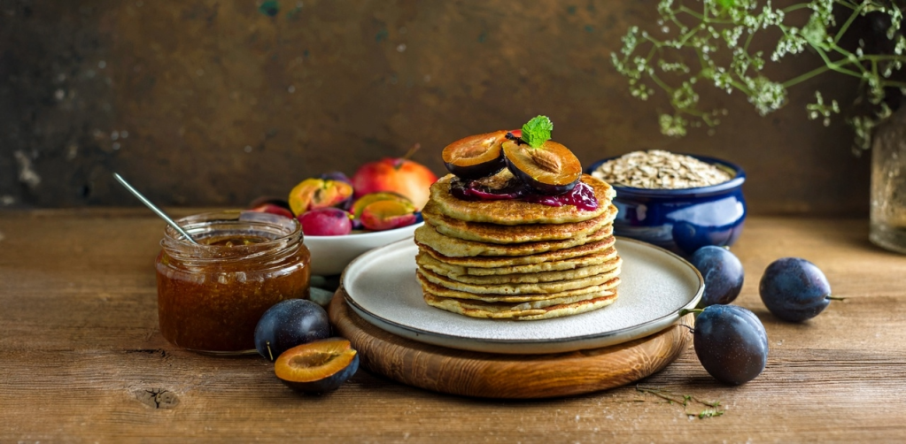A stack of thin Palatschinken pancakes with powidl plum jam on a wooden board, surrounded by fresh plums, buckwheat grains, and herbs, reflecting a sustainable Bohemian Forest theme.
