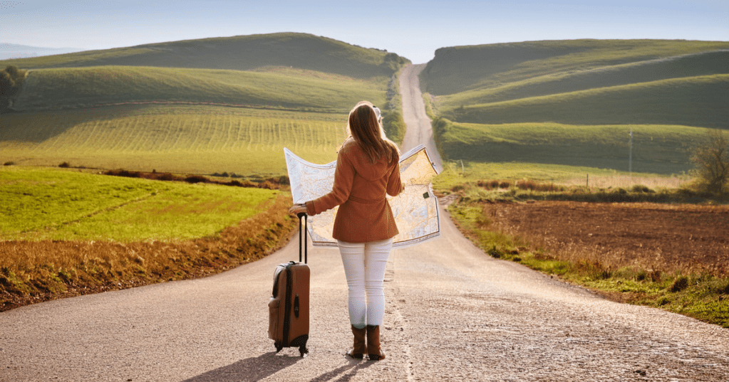 Ein Mensch steht an einer Kreuzung beim Auswandern in einer ruhigen Landschaft, mit einem abgenutzten Koffer in der einen und einer offenen Karte in der anderen Hand.