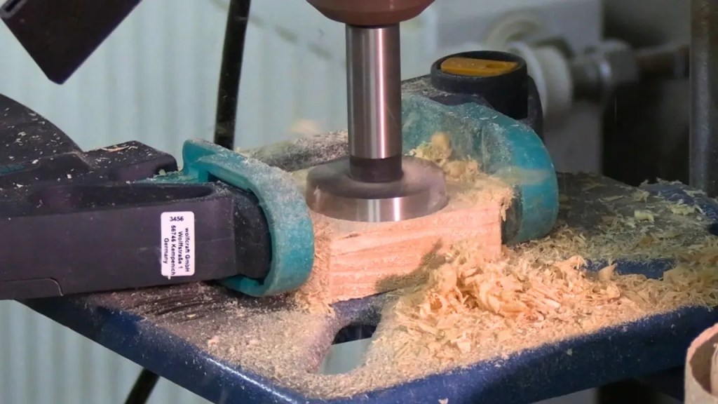 Ein Forstnerbohrer in einer Ständerbohrmaschine bohrt ein Loch in ein Stück Holzbrett. Gehalten von einer Schraubzwinge