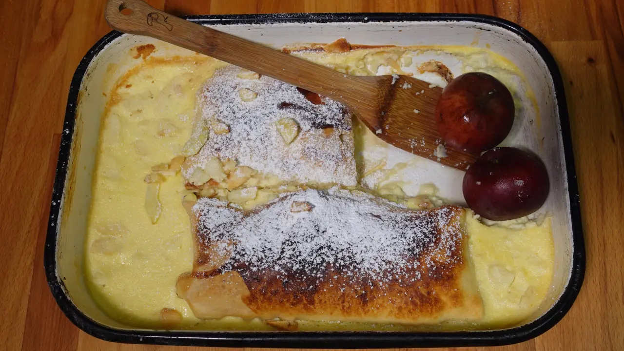 Ein frisch gebackener, goldbrauner Apfel-Milchrahmstrudel in einer klassischen weißen Emailleform mit schwarzem Rand. Der Strudel ist mit Puderzucker bestäubt und liegt in einem gestockten Milchguss, dekoriert mit zwei ganzen roten Äpfeln und einem hölzernen Pfannenwender